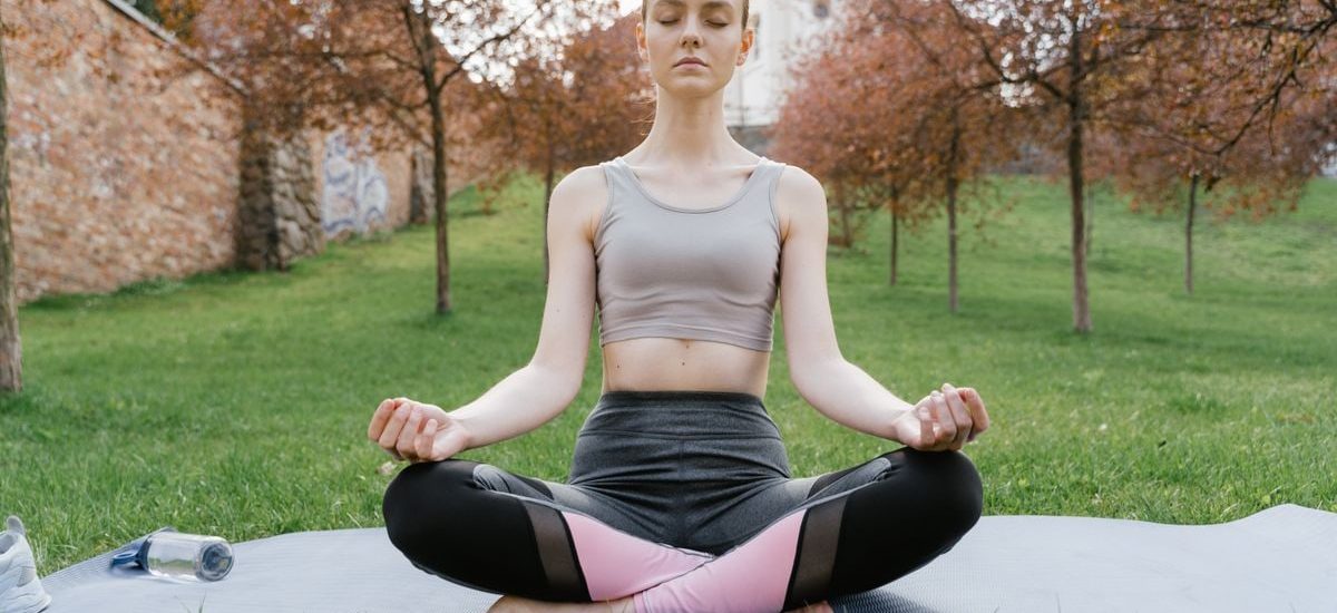 A woman performs meditation in a serene park setting on a yoga mat.