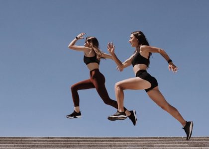 Two women in active wear leap energetically on stairs against a clear blue sky.